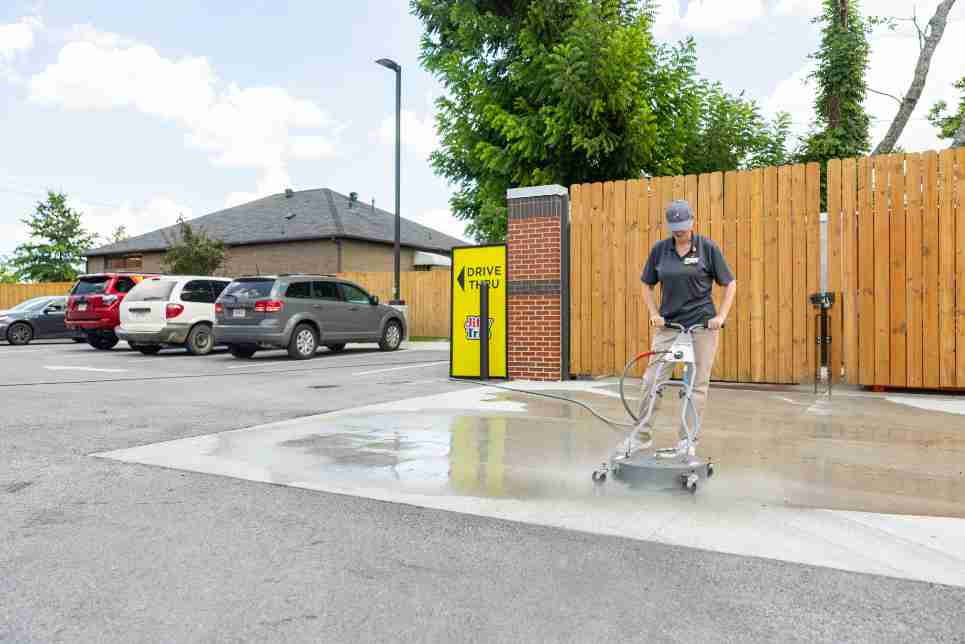 Convenience store employee using Spraymaster pressure washing system to clean drive-thru concrete, reducing labor time and improving exterior appearance